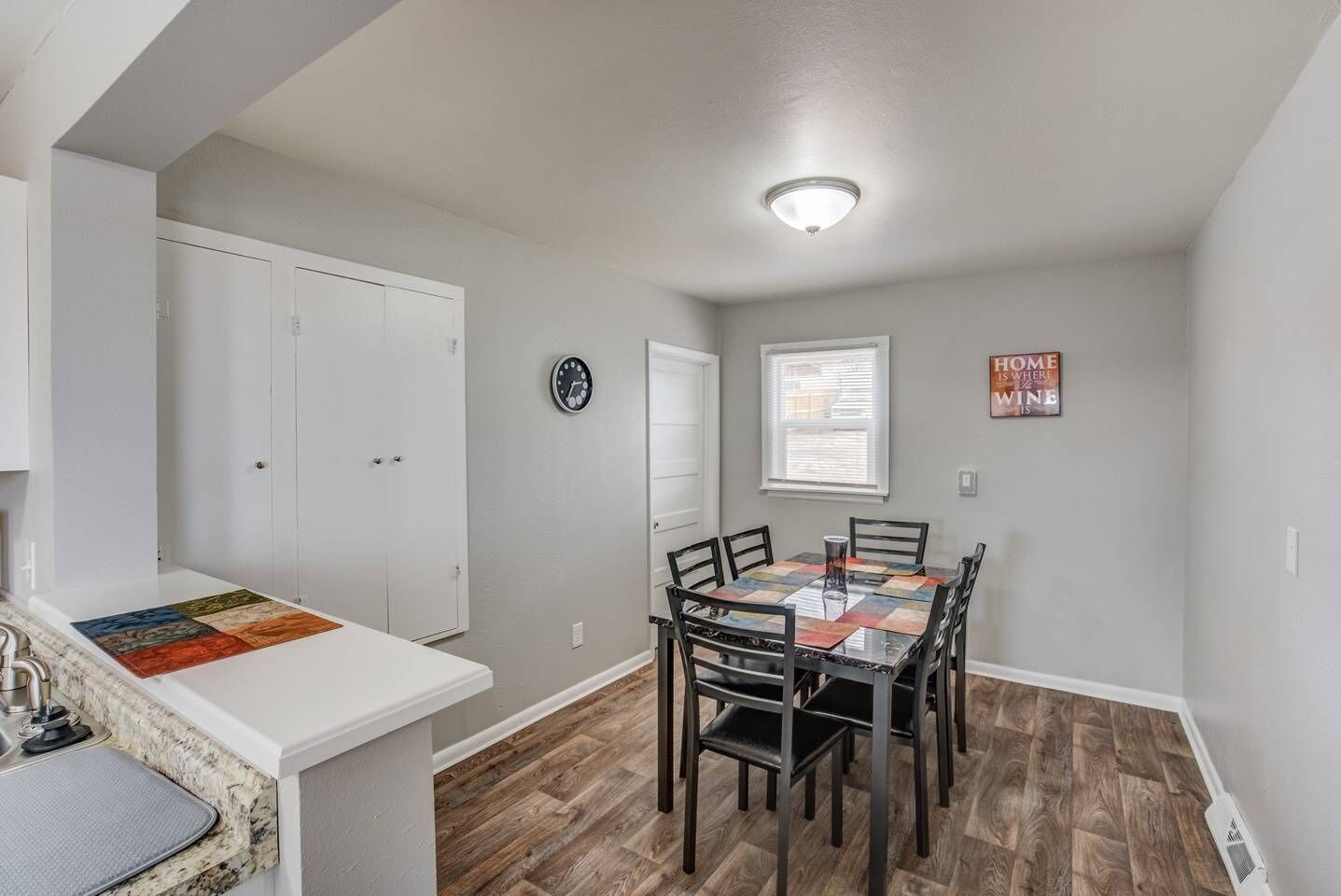 A dining area with a table and chairs, a clock on the wall, and a sign that says "HOME WHERE VINE IS".