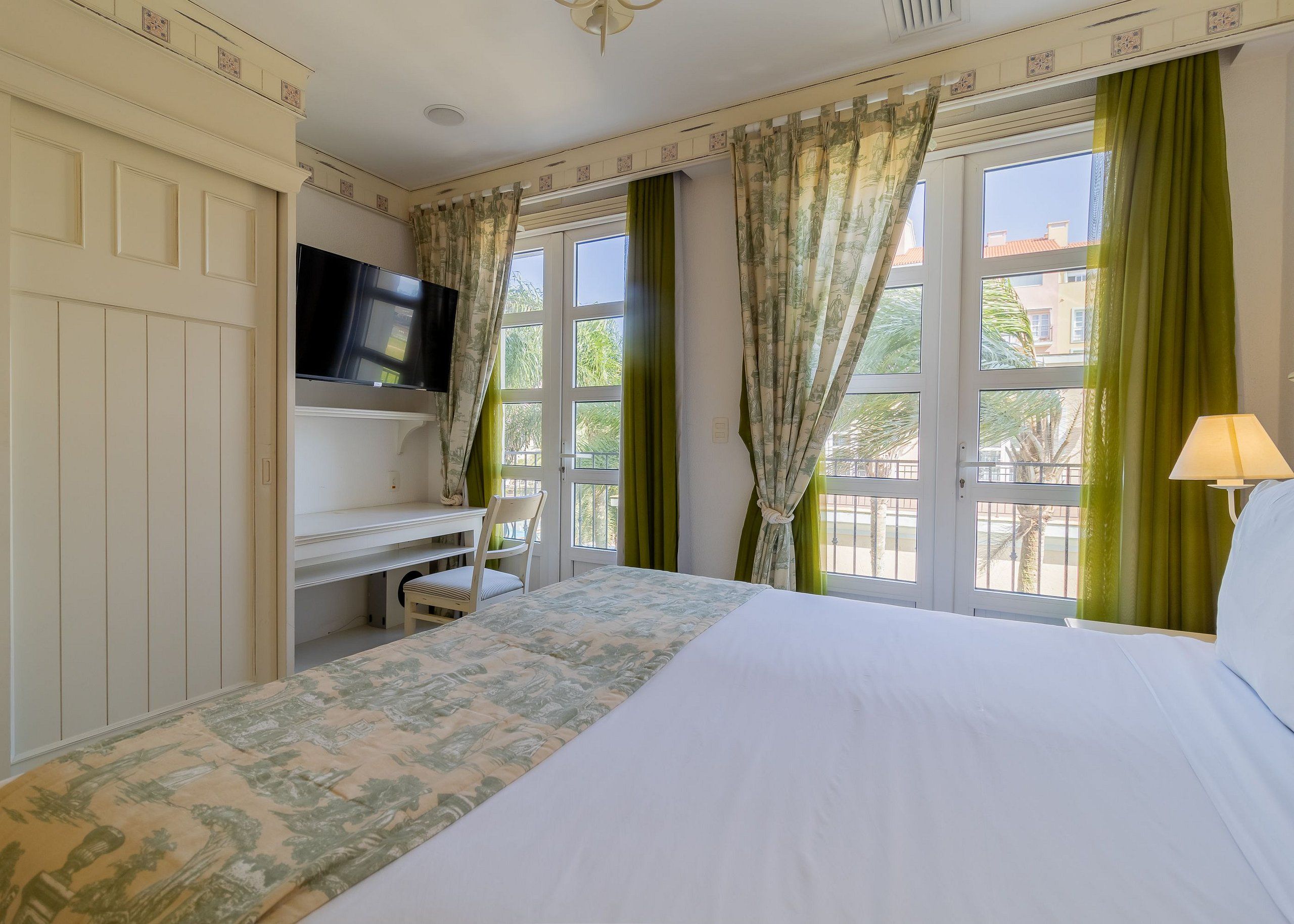 Quarto de hotel com cama, escrivaninha, televisão e janela com cortinas.