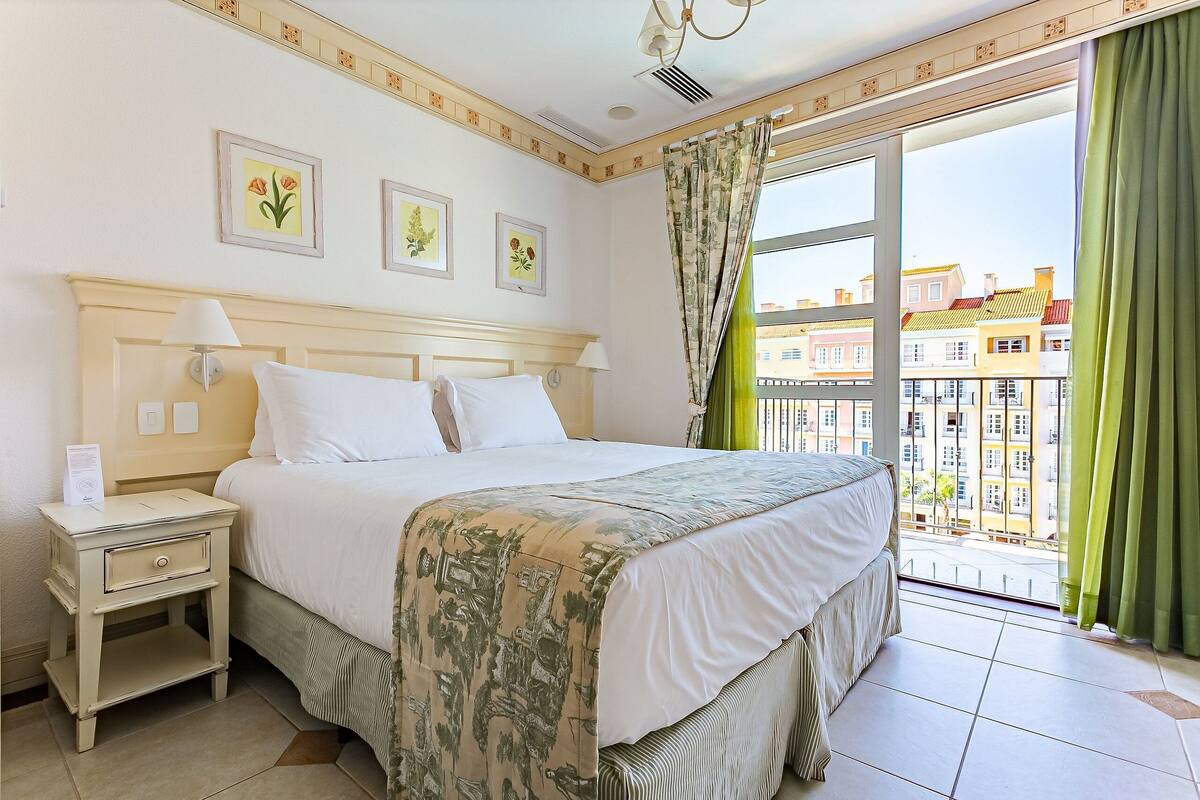 Habitación de hotel con cama, mesita de noche y ventana con vista a edificios.