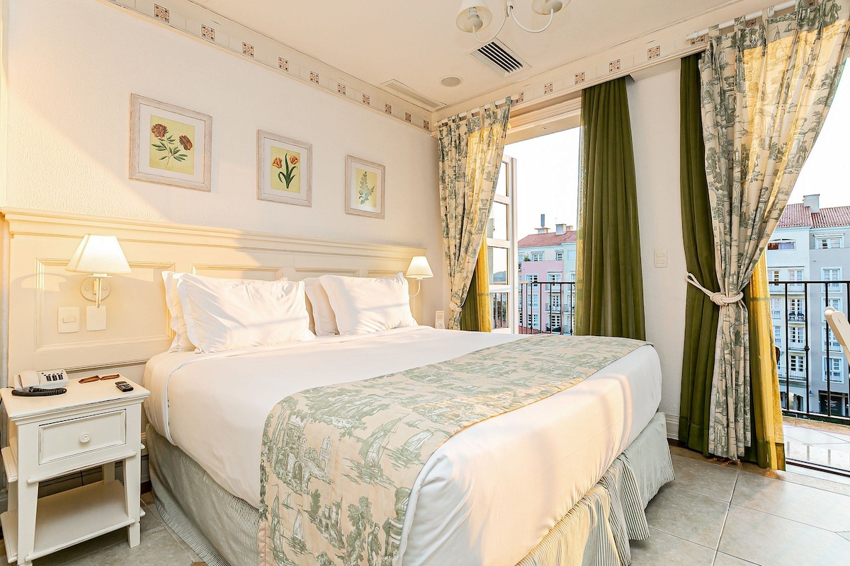 Habitación de hotel con una cama grande, dos mesitas de noche, una ventana con cortinas y un cuadro con motivos florales en la pared.