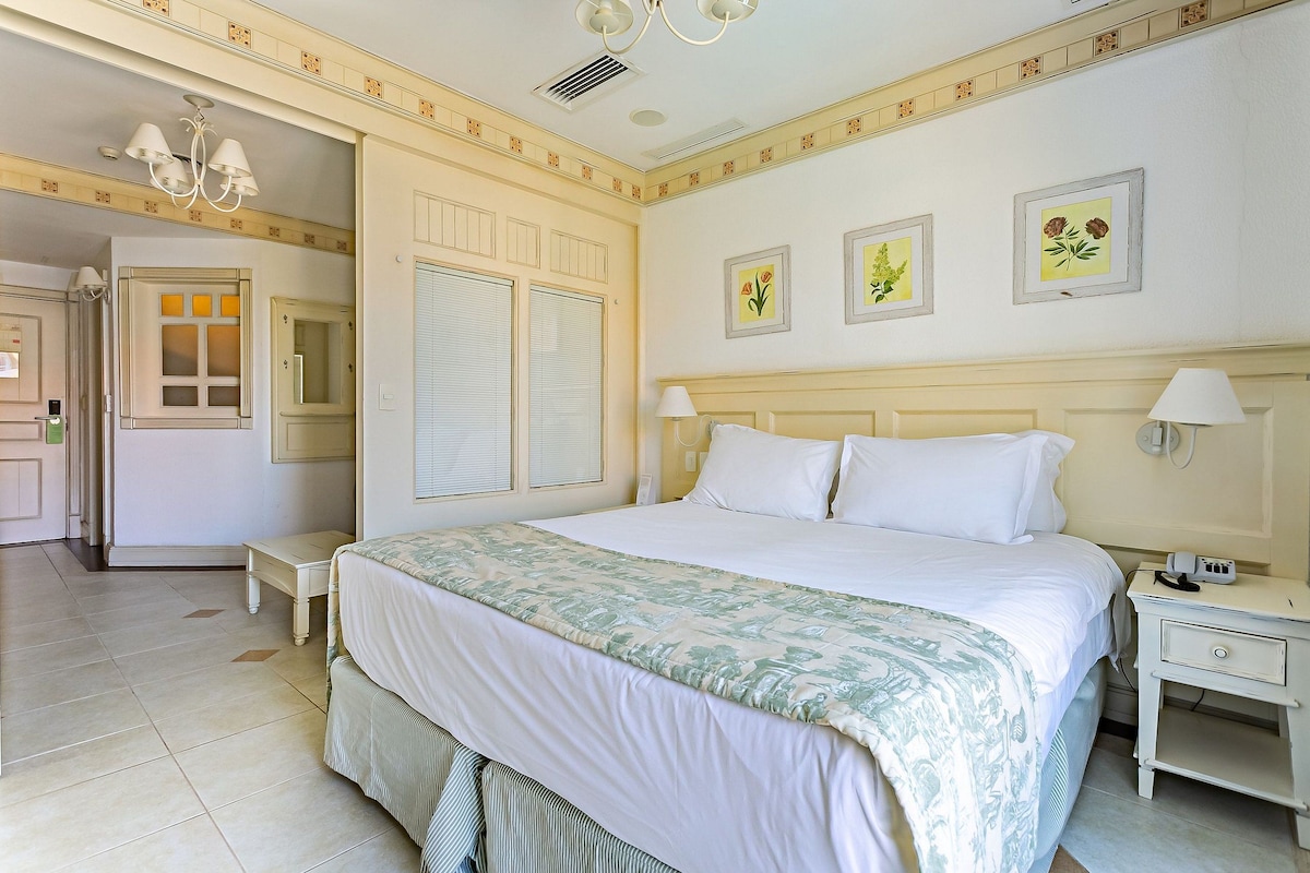 Habitación de hotel con cama, mesitas de noche, lámpara de noche, candelabro e impresiones botánicas enmarcadas en la pared.
