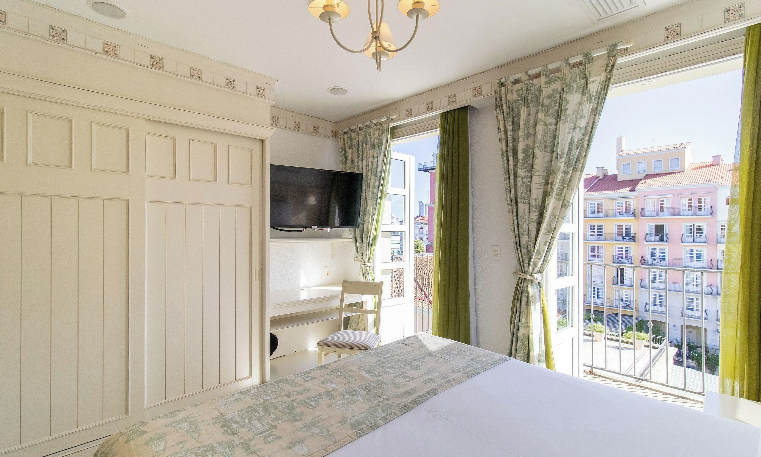 Ein Hotelzimmer mit Bett, Fernseher, Sessel und Blick auf Gebäude durch das Fenster.