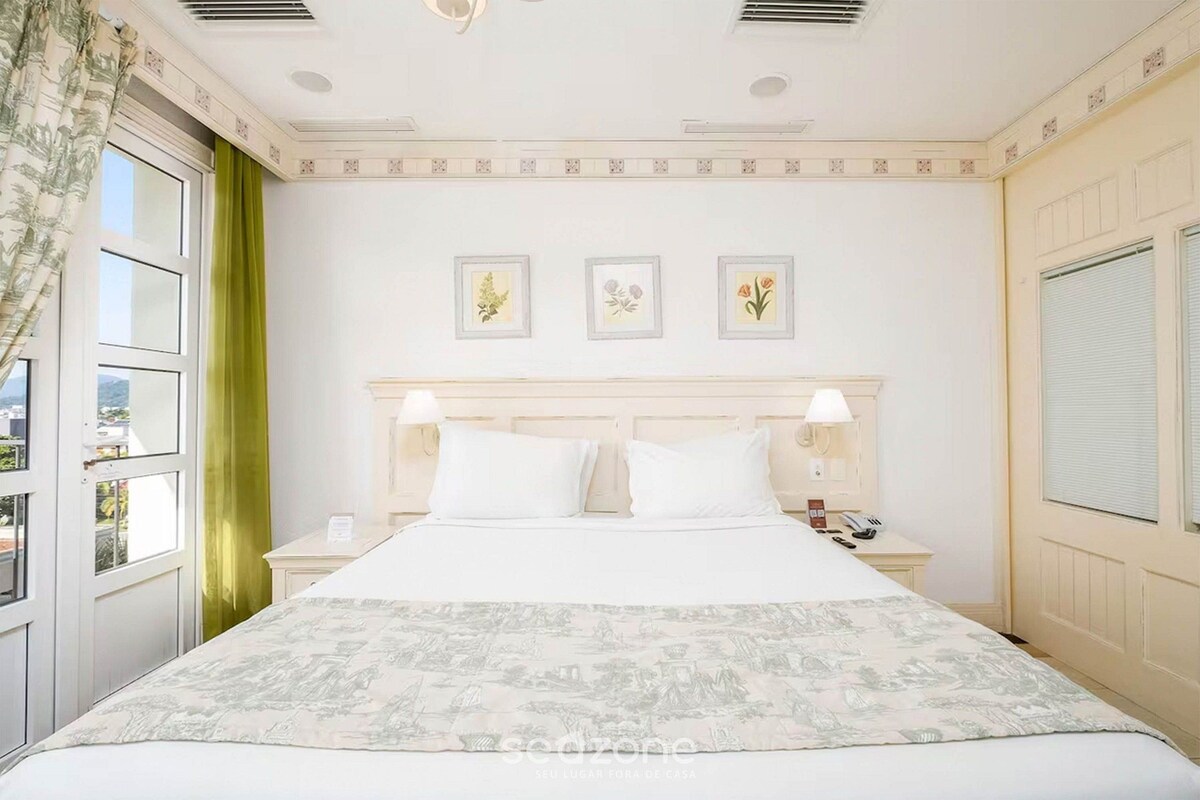 Una habitación de hotel con una cama, dos mesitas de noche, una ventana con cortinas y tres cuadros en la pared.