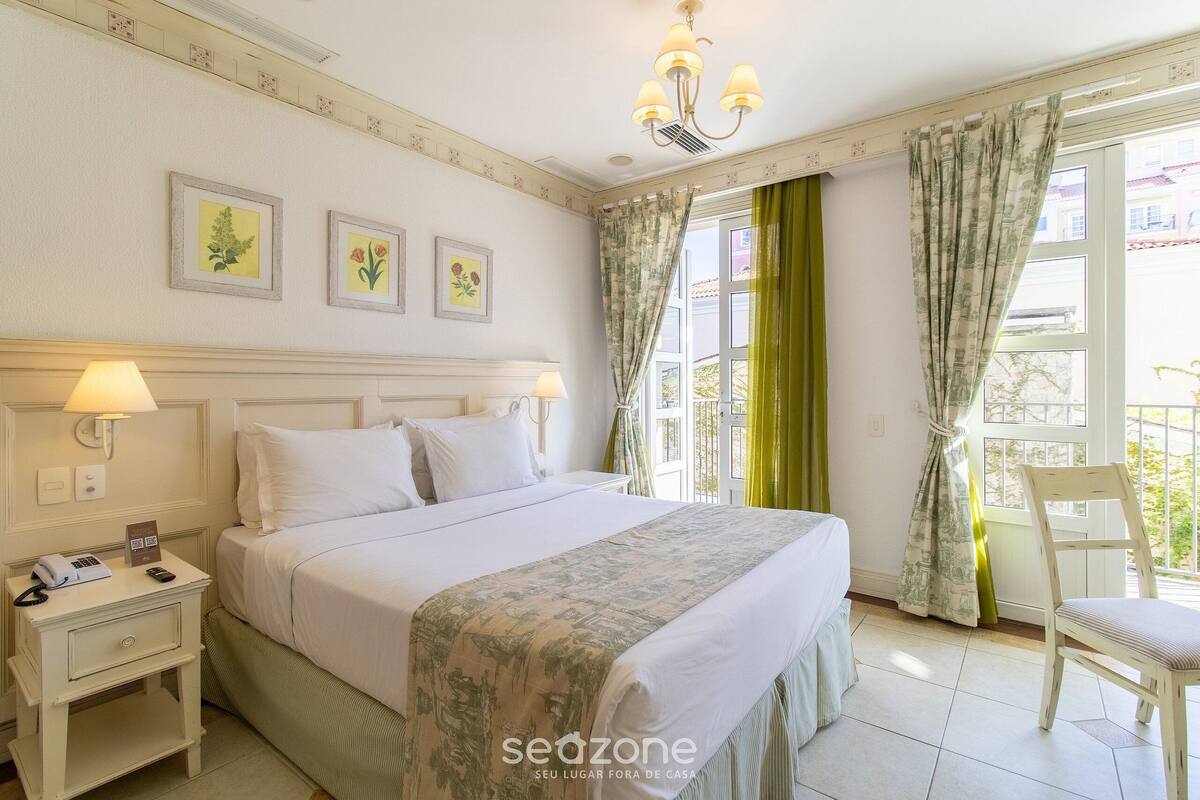 Habitación de hotel con cama, lámparas de noche, mesita de noche, una silla y una ventana con cortinas.