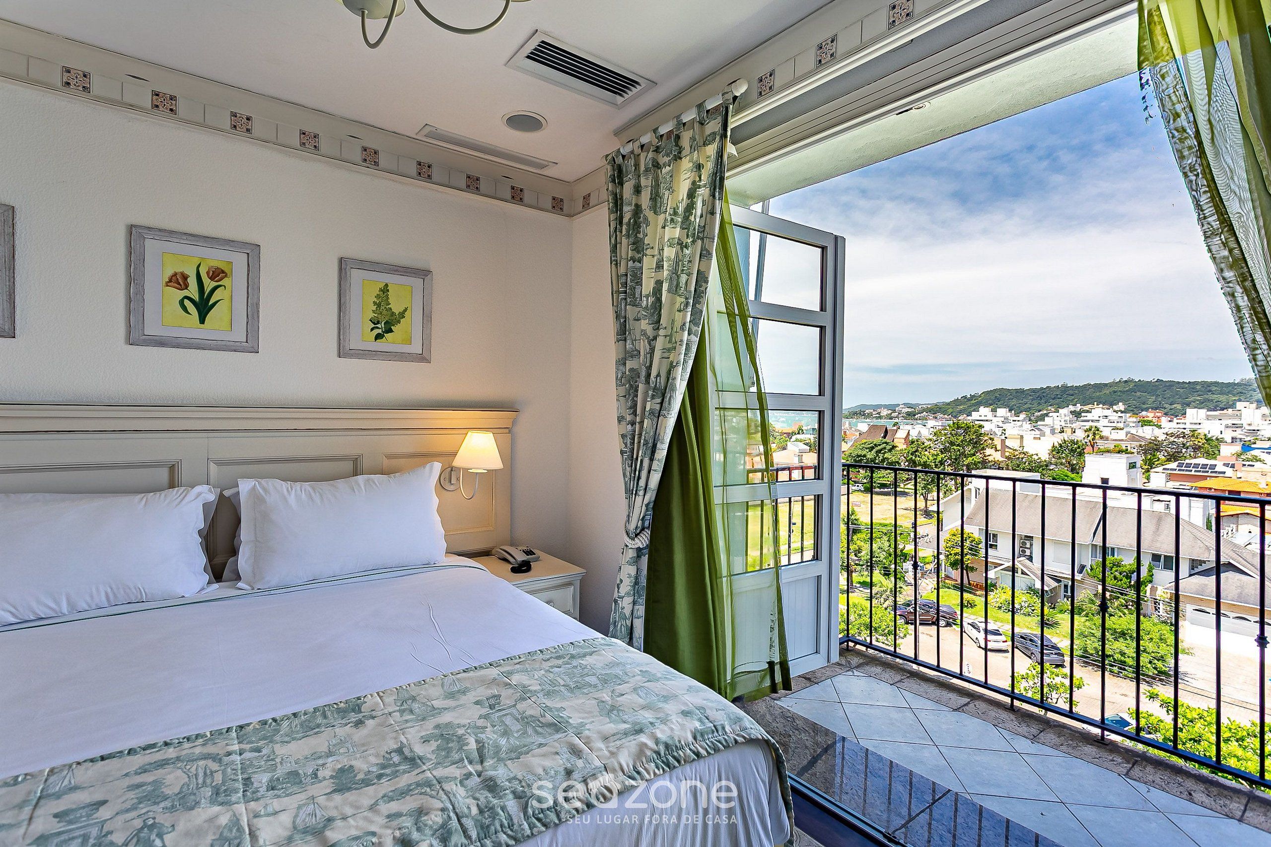 Habitación de hotel con cama, mesita de noche, lámpara, dos cuadros enmarcados y balcón con vistas a la ciudad.