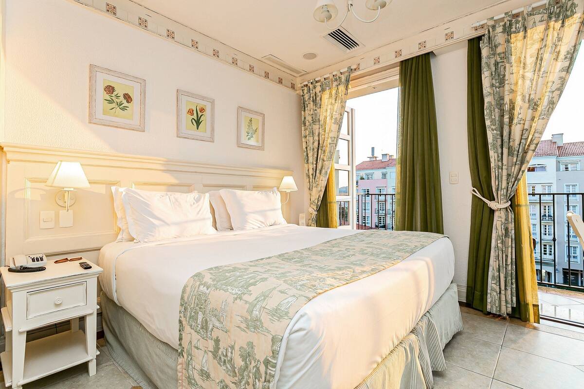 Habitación de hotel con una cama grande, dos mesitas de noche, una ventana con cortinas y un cuadro con motivos florales en la pared.