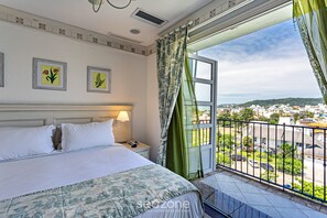 A hotel room with a bed, a nightstand, a lamp, two framed pictures, and a balcony with a view of the city. - Confortáveis Studios no IL Campanario Resort ILCJR (Florianópolis)