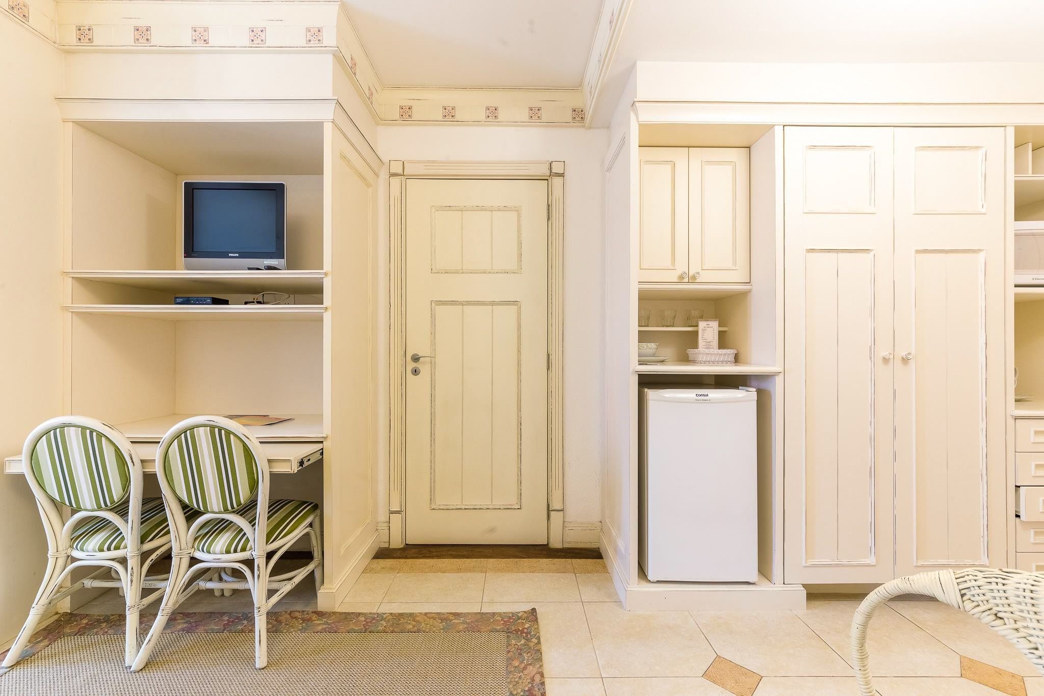 Ein kleines Zimmer mit Schreibtisch, zwei Stühlen, Fernseher, Kleiderschrank und Kühlschrank.