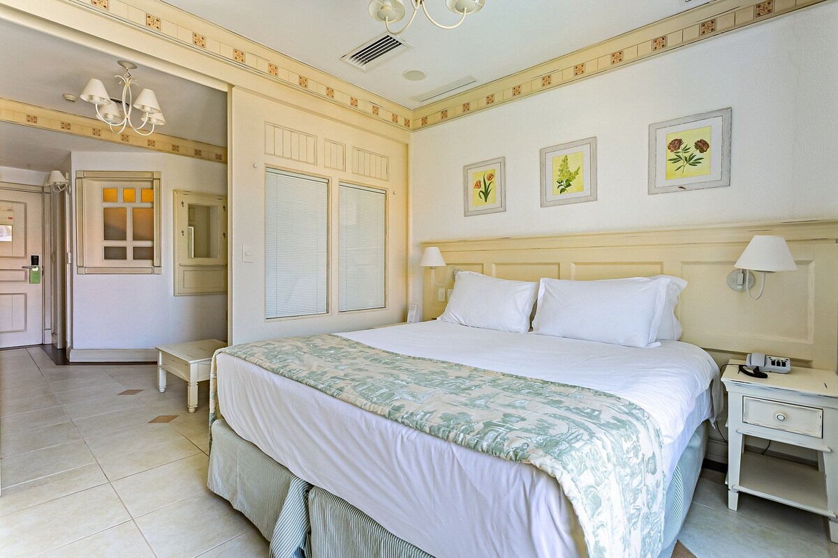 Habitación de hotel con cama, mesitas de noche, lámpara de noche, candelabro e impresiones botánicas enmarcadas en la pared.