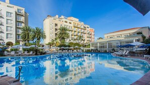 Una piscina con sillones de descanso y sombrillas, rodeada de palmeras y edificios de hotel.