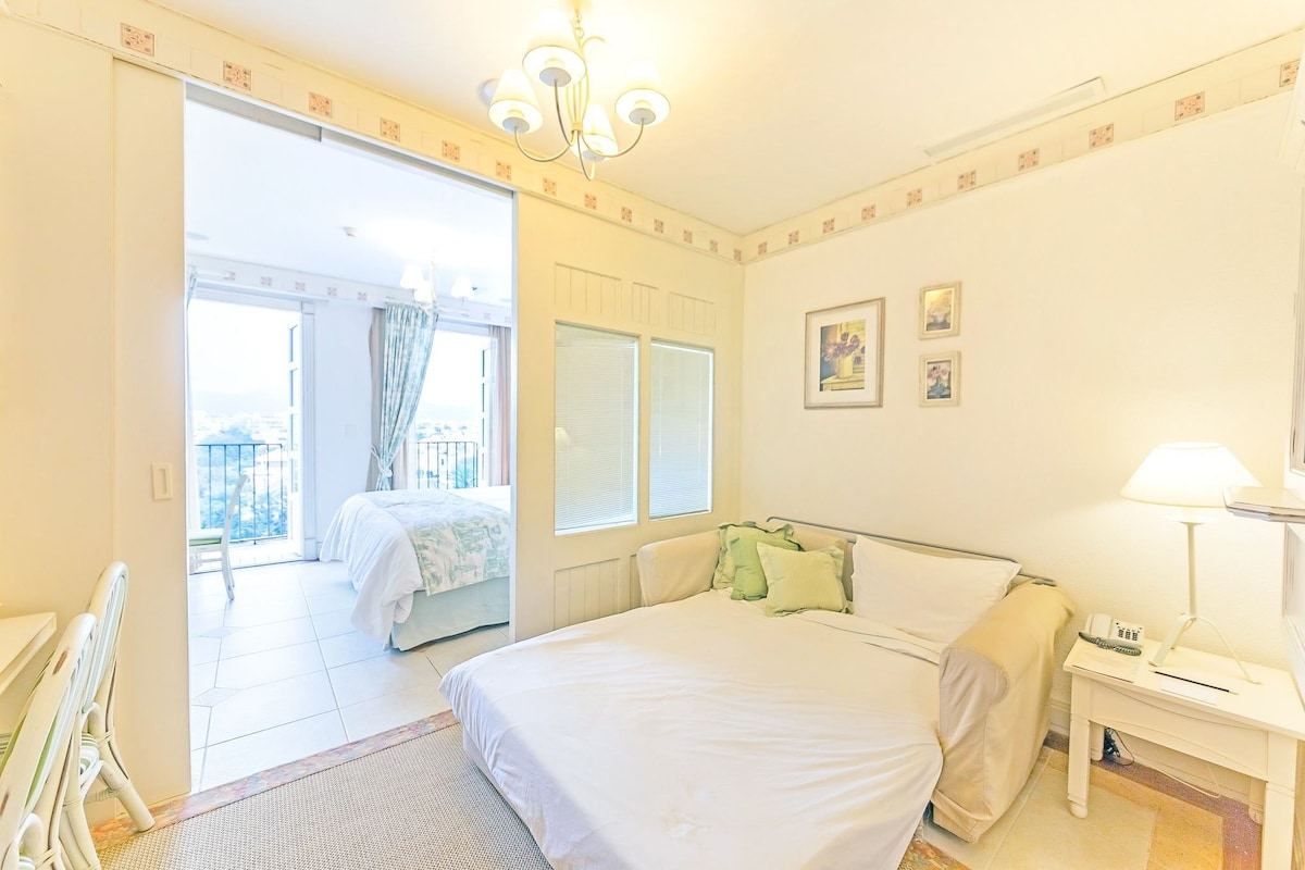 Habitación de hotel con cama, mesita de noche con lámpara, una silla, una ventana y puerta que da a un balcón.