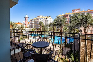 A balcony with a view of a swimming pool, palm trees, and buildings in the background.