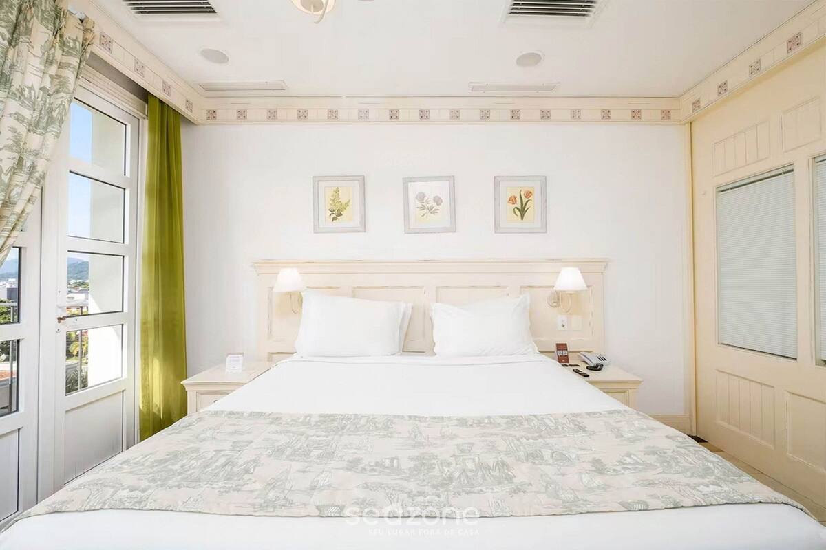 Una habitación de hotel con una cama, dos mesitas de noche, una ventana con cortinas y tres cuadros en la pared.