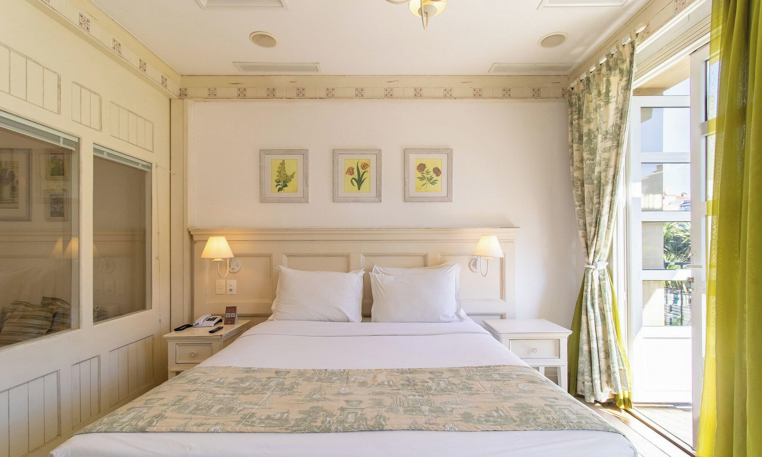 Quarto de hotel com uma cama grande, duas mesas de cabeceira, um criado-mudo, uma janela com cortinas e três impressões botânicas emolduradas na parede.
