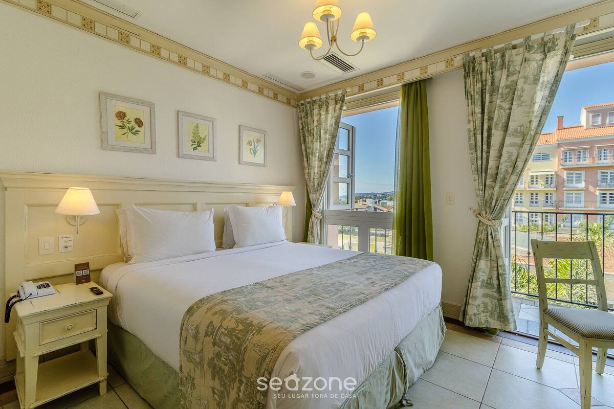 A hotel room with a bed, bedside lamps, a nightstand, a chair, a window with curtains, and a balcony view.