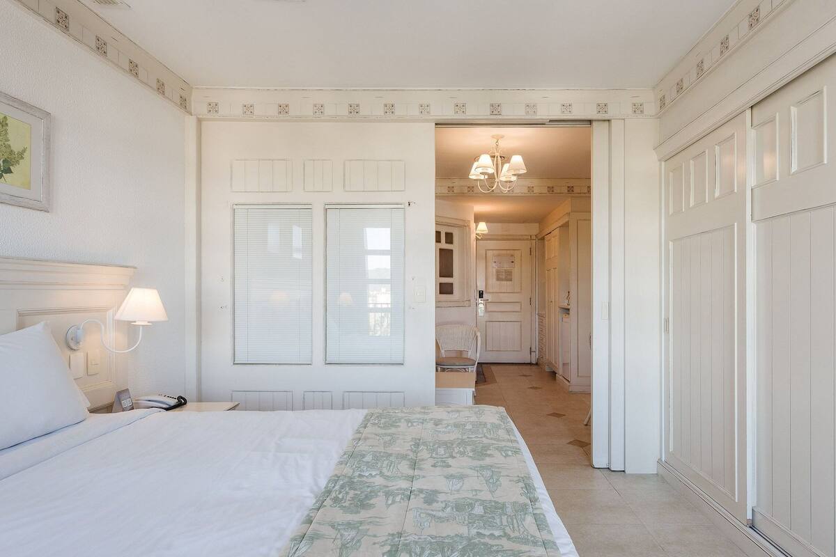 A hotel room with a bed, bedside lamps, a window with blinds, a door leading to a hallway, and a chandelier in the hallway.