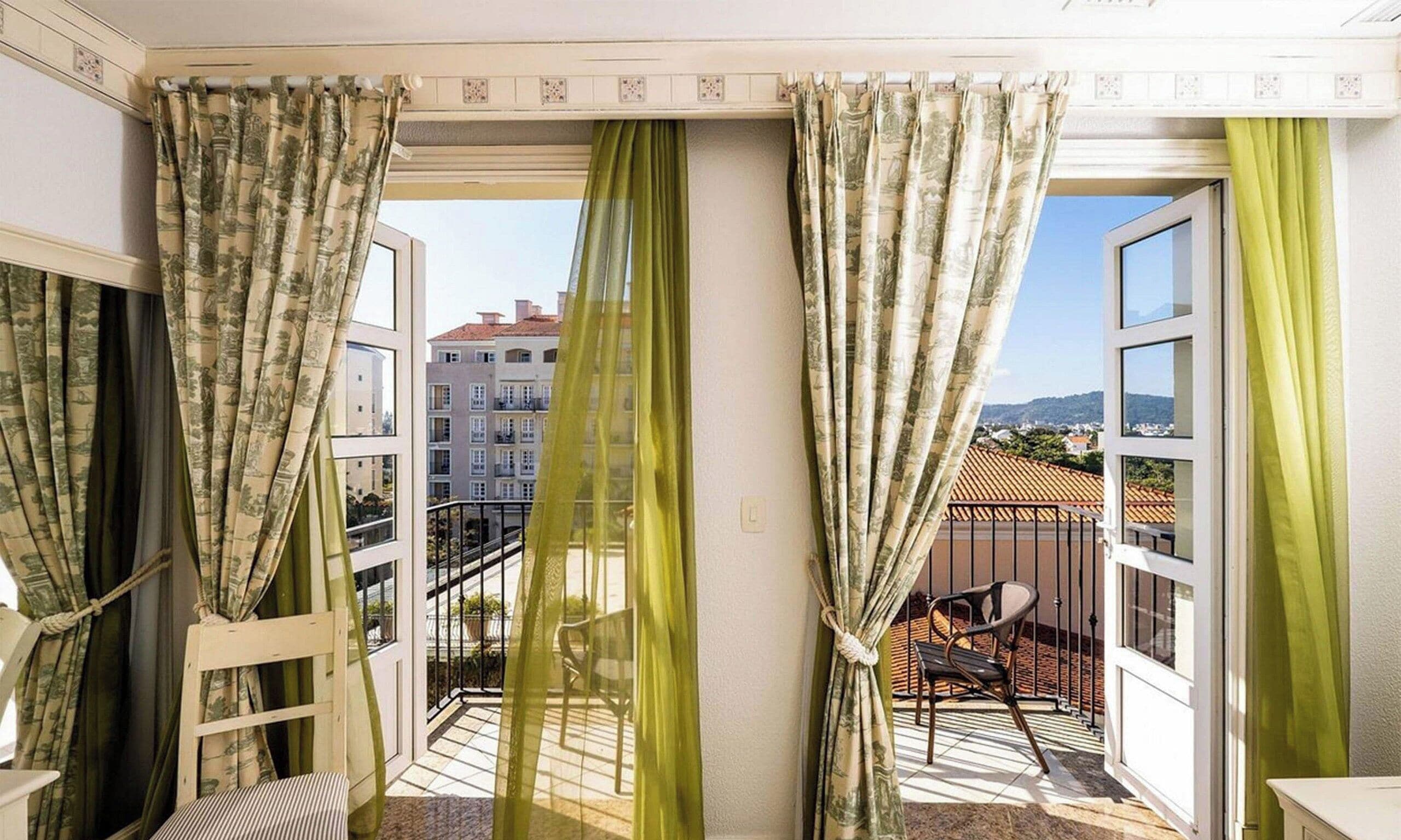 A balcony with a view of buildings, a chair, and curtains.