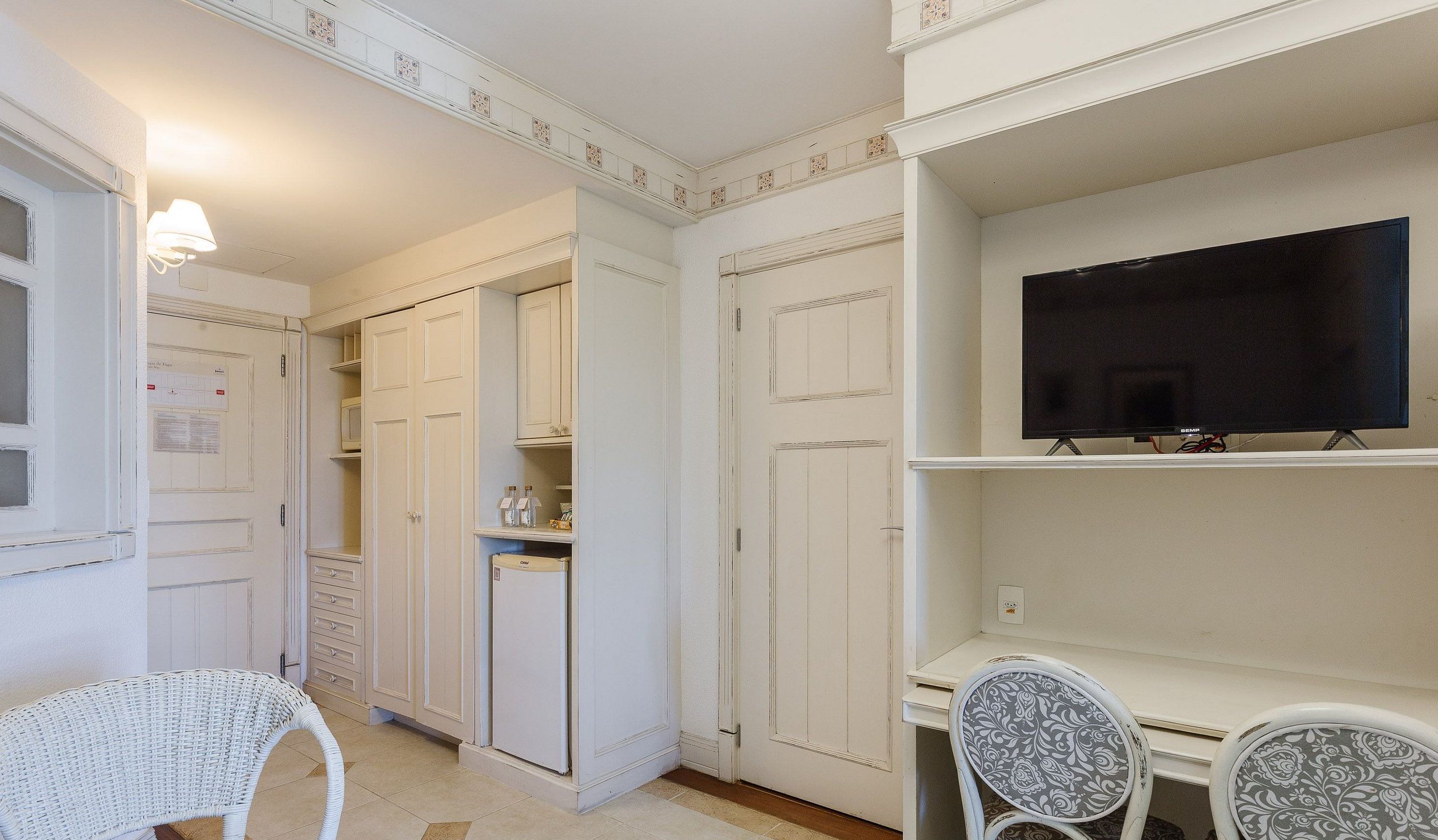 A room with a white wardrobe, a small refrigerator, a television on a stand, and a wicker chair.
