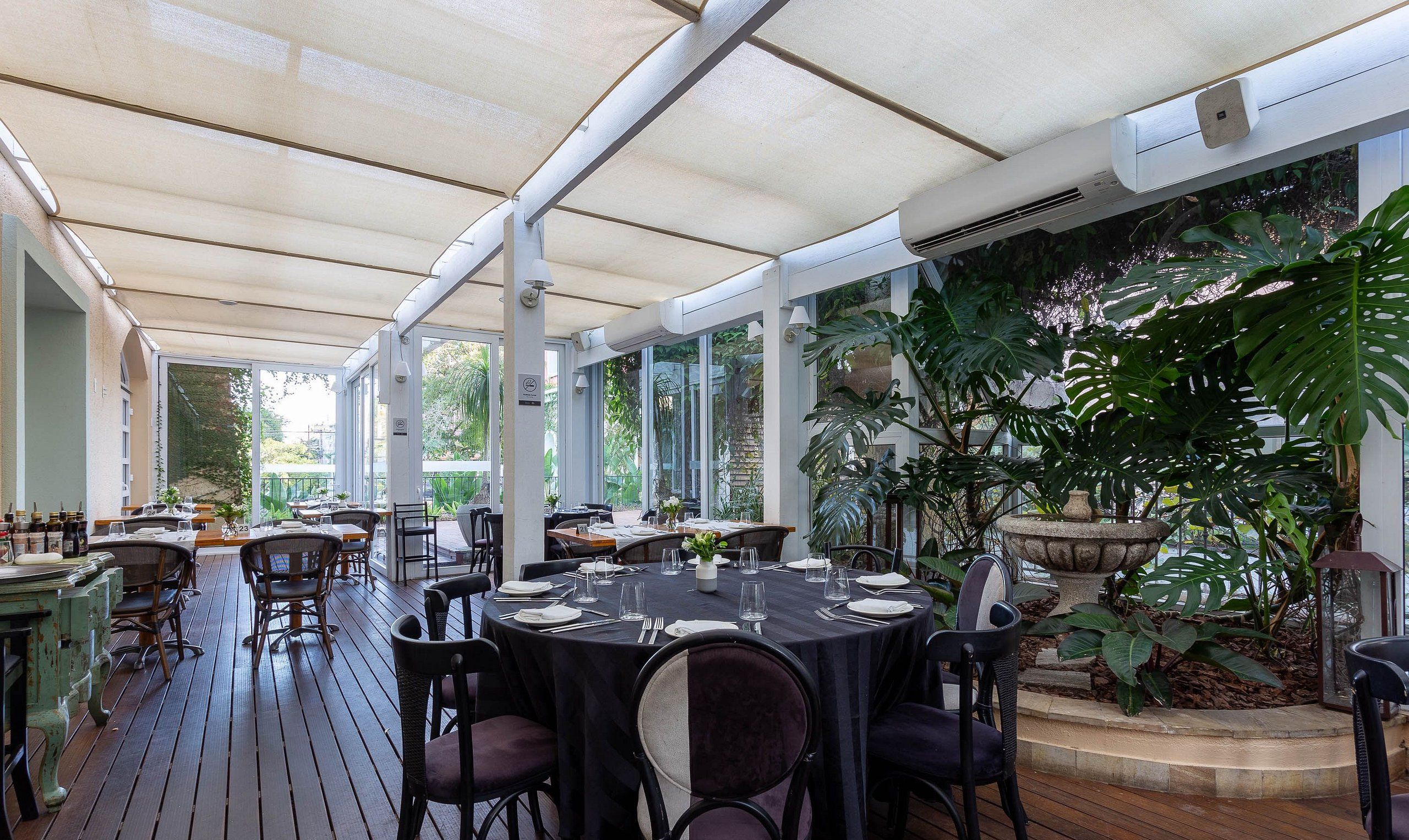 An outdoor dining area with round tables and chairs, covered by a translucent roof, surrounded by large windows and potted plants.