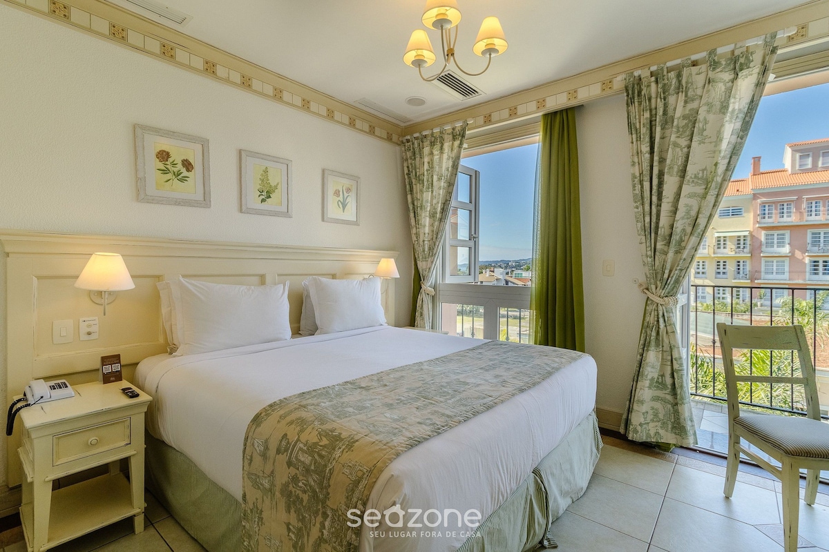 Habitación de hotel con cama, lámparas de noche, mesita de noche, una silla, ventana con cortinas y vistas al balcón.
