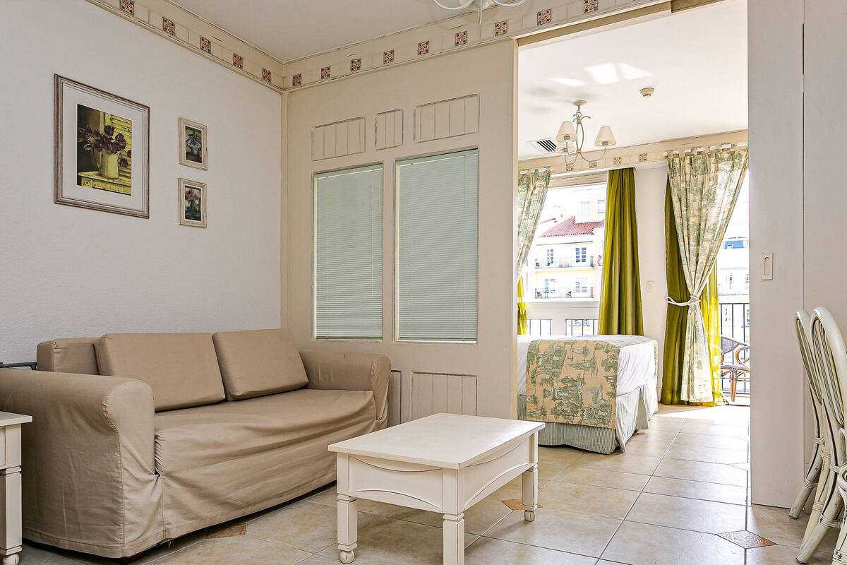 Un salón con sofá, una mesita y un radiador. Hay cuadros enmarcados en la pared, una ventana con persianas y vistas al exterior.