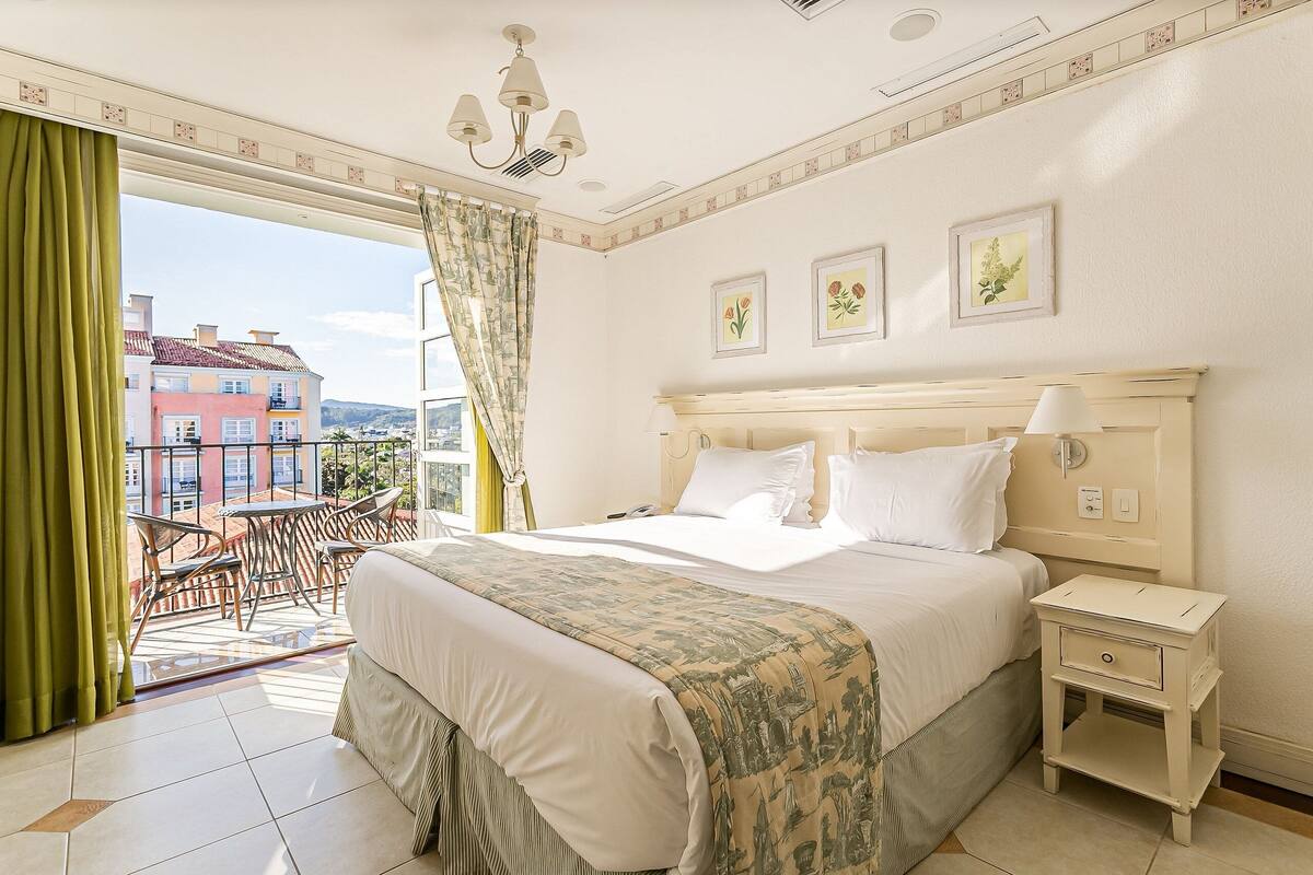 Habitación de hotel con cama, mesita de noche, una silla y vista a edificios a través de una ventana.