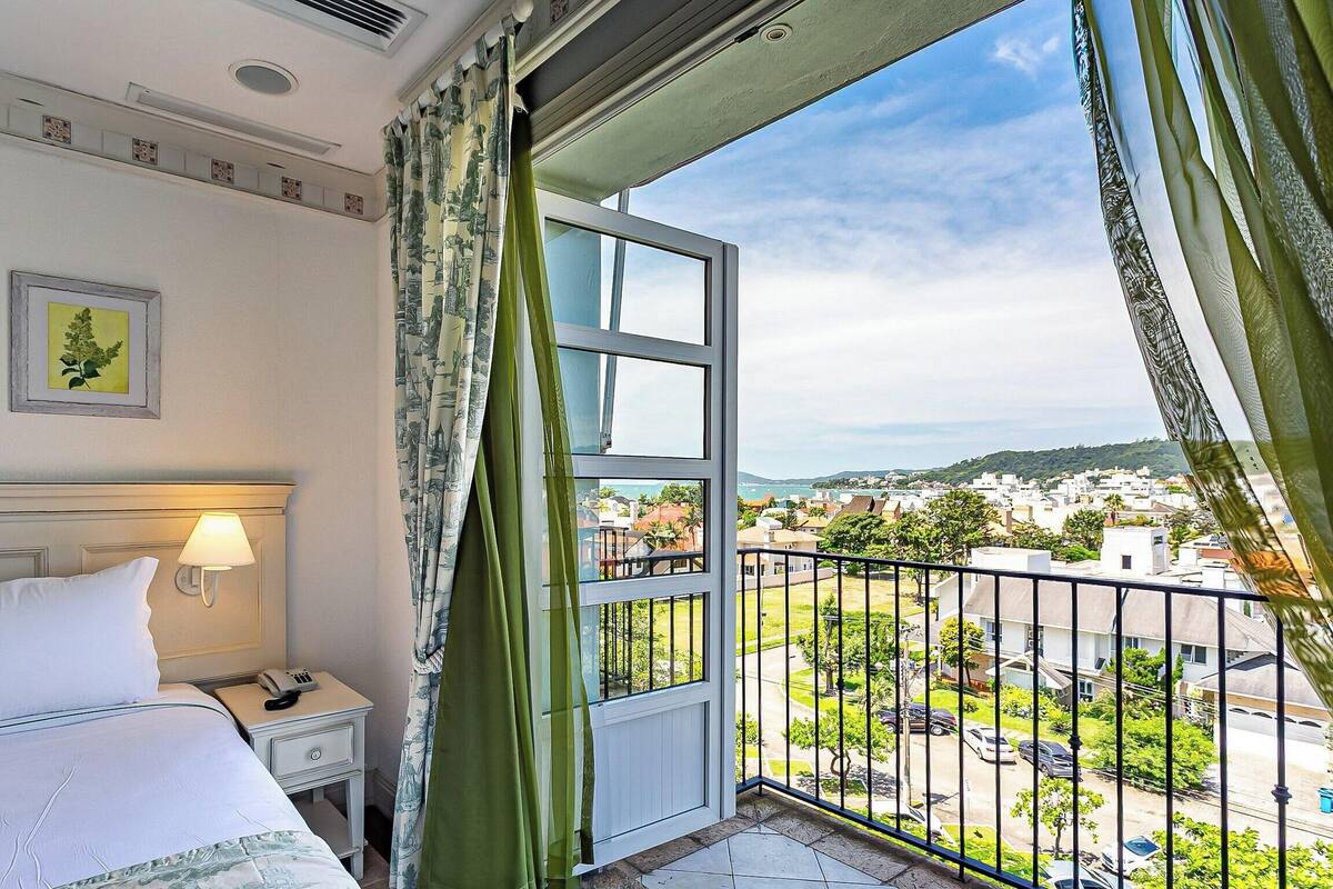 Habitación de hotel con cama, mesita de noche con lámpara, teléfono y ventana con vistas a un pueblo y colinas.