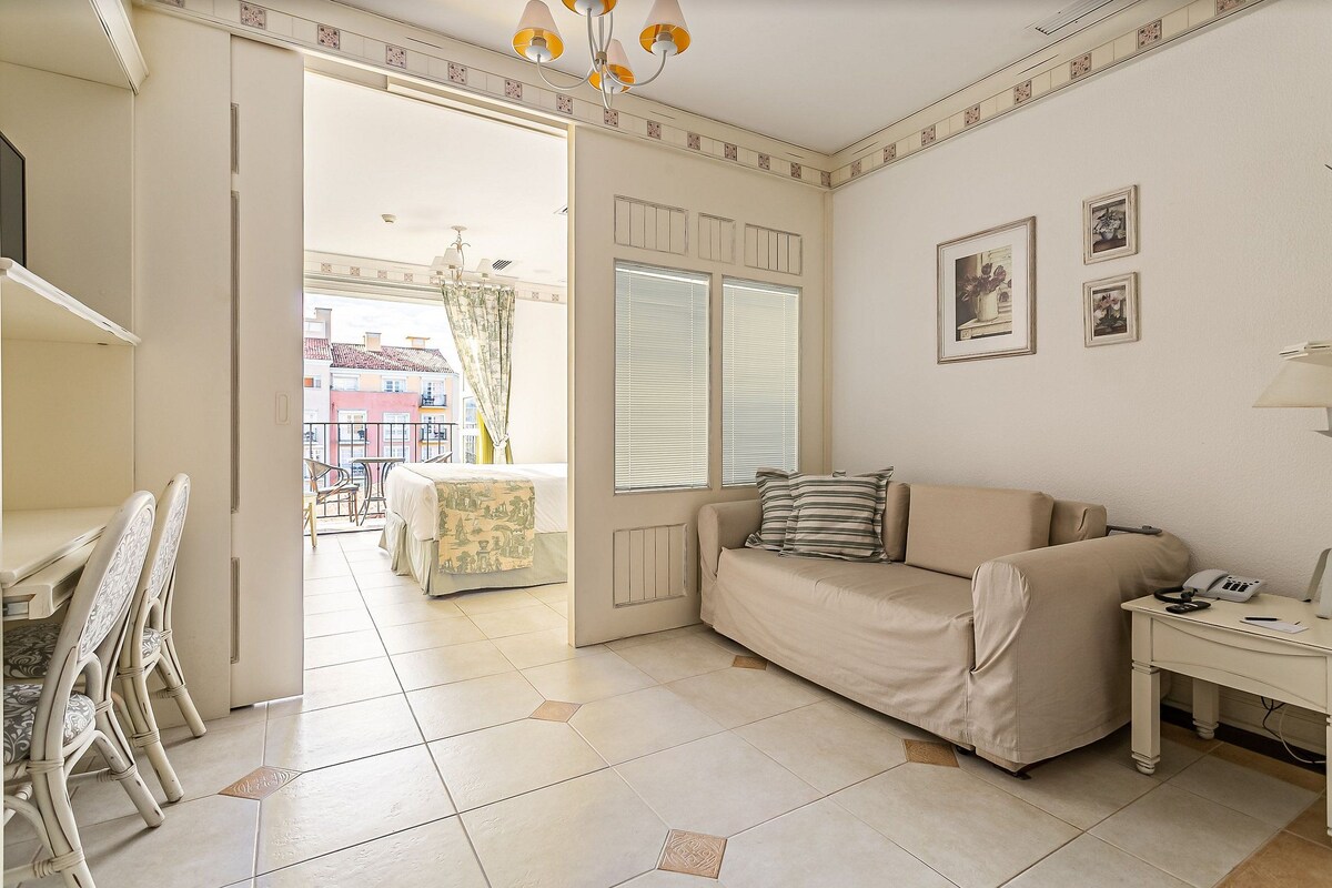 Una sala de estar con un sofá, una mesa de comedor con sillas, una mesita con una lámpara y una ventana con persianas.