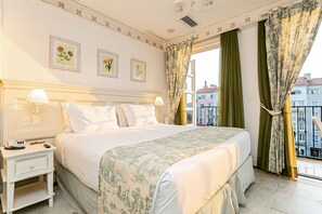 Habitación de hotel con una cama grande, dos mesitas de noche, una ventana con cortinas y un cuadro con motivos florales en la pared.