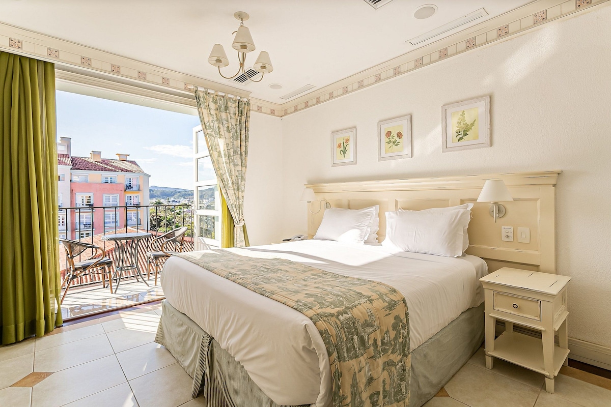 Habitación de hotel con cama, mesita de noche, una silla y vista a edificios a través de una ventana.