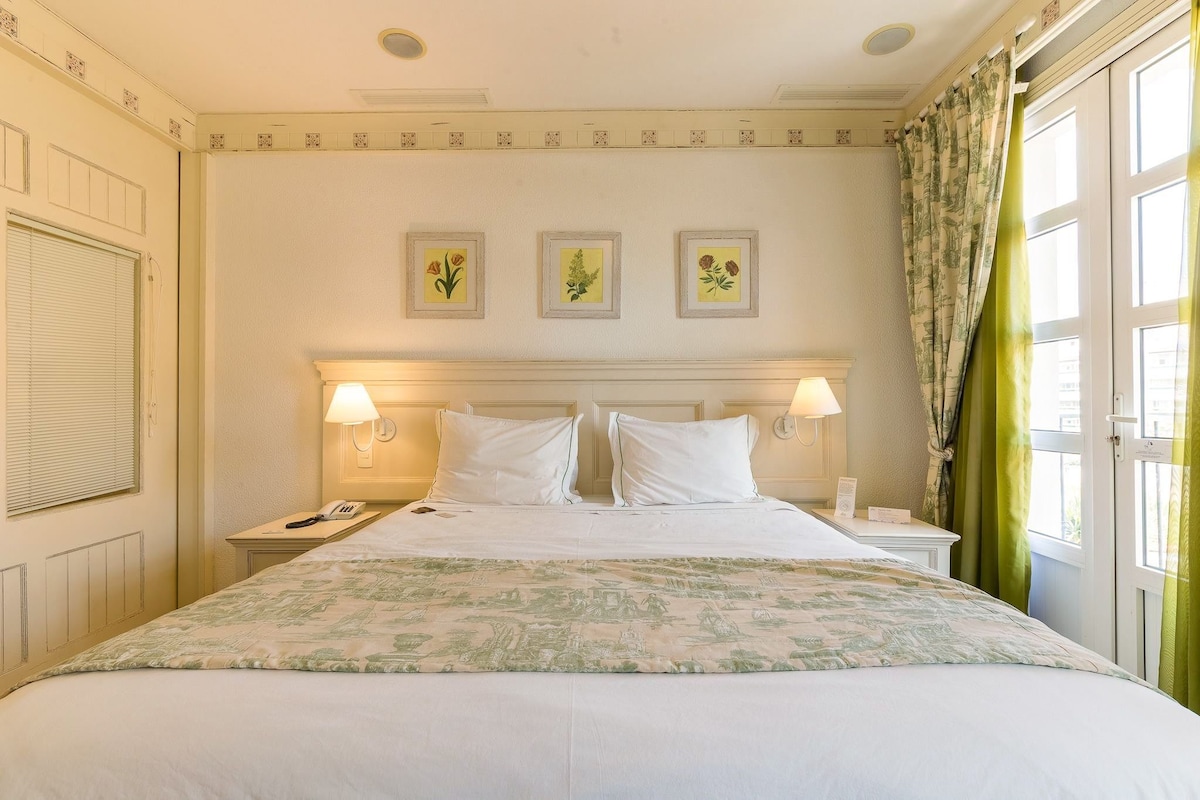 Una habitación de hotel con cama, lámparas de noche, teléfono y ventana con cortinas.