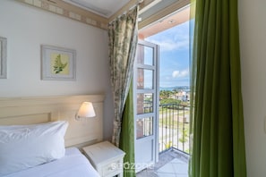 Habitación de hotel con cama, mesita de noche, lámpara de pared, un cuadro enmarcado y balcón con vistas.