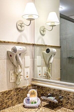 Un baño con dos secadores de pelo, lámparas de pared, un espejo y una cesta de artículos de aseo.