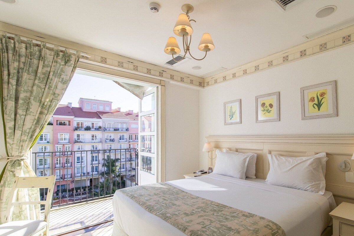 A hotel room with a bed, a nightstand, a lamp, a window with curtains, and a view of buildings.