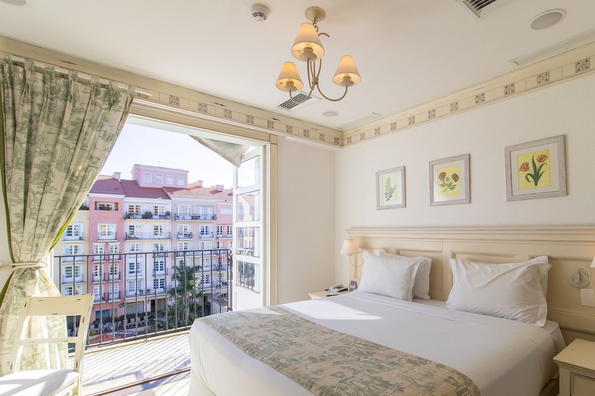 Habitación de hotel con cama, mesita de noche, lámpara, ventana con cortinas y vista a edificios.