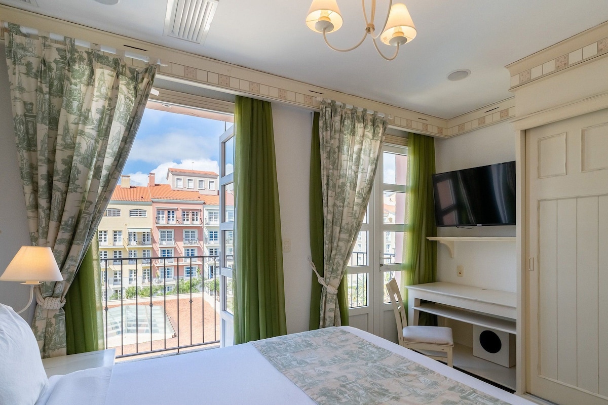 A hotel room with a bed, a desk, a TV, and a balcony with curtains.