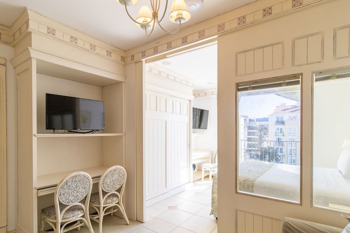Una habitación con televisión en un estante, dos sillones estampados, una cama con vistas a edificios y un balcón.