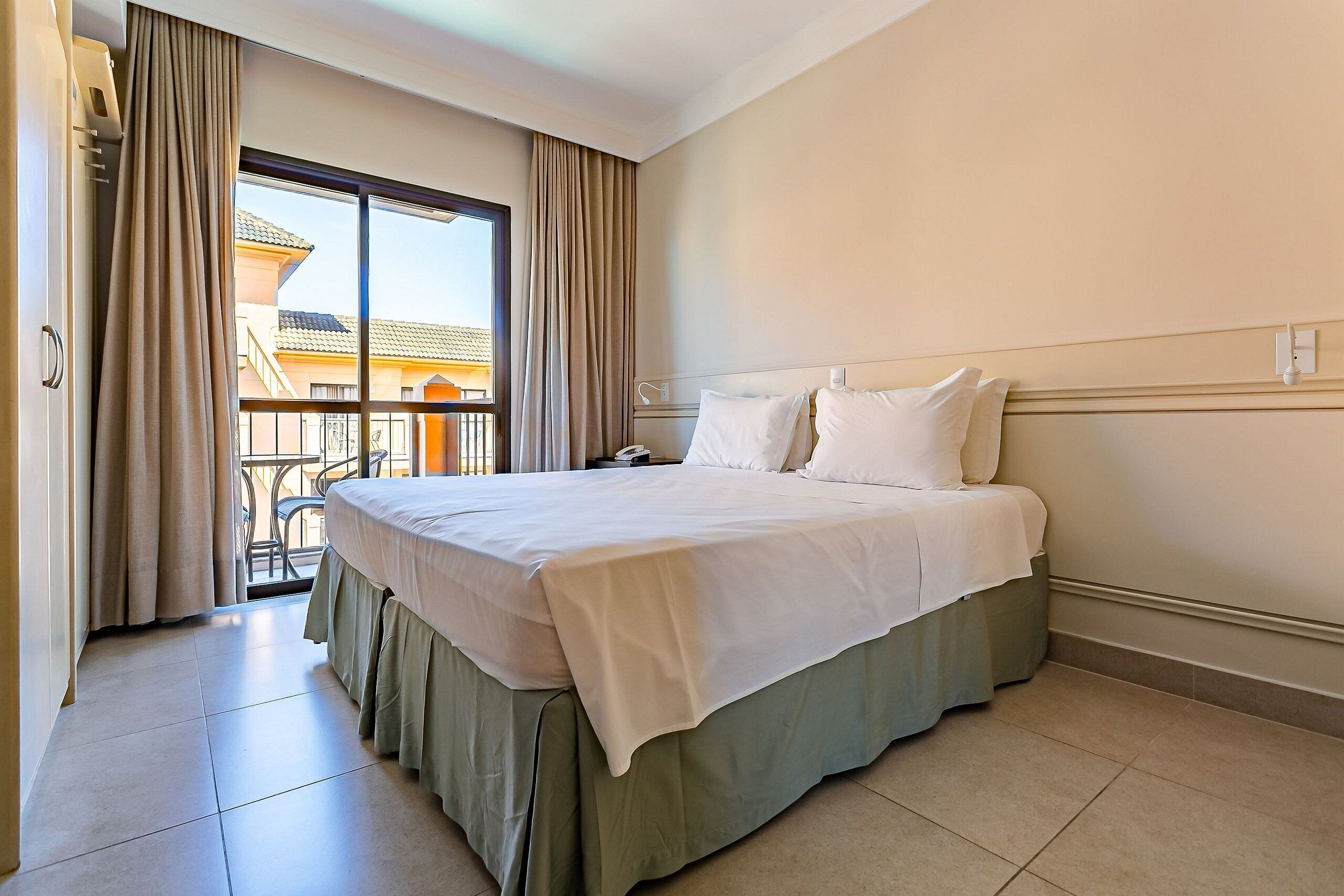Habitación de hotel con una cama grande, un balcón con cortinas y vistas a edificios.