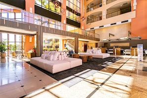 A spacious lobby with a high ceiling, a central seating area with sofas, a reception desk, and a staircase leading to upper floors.