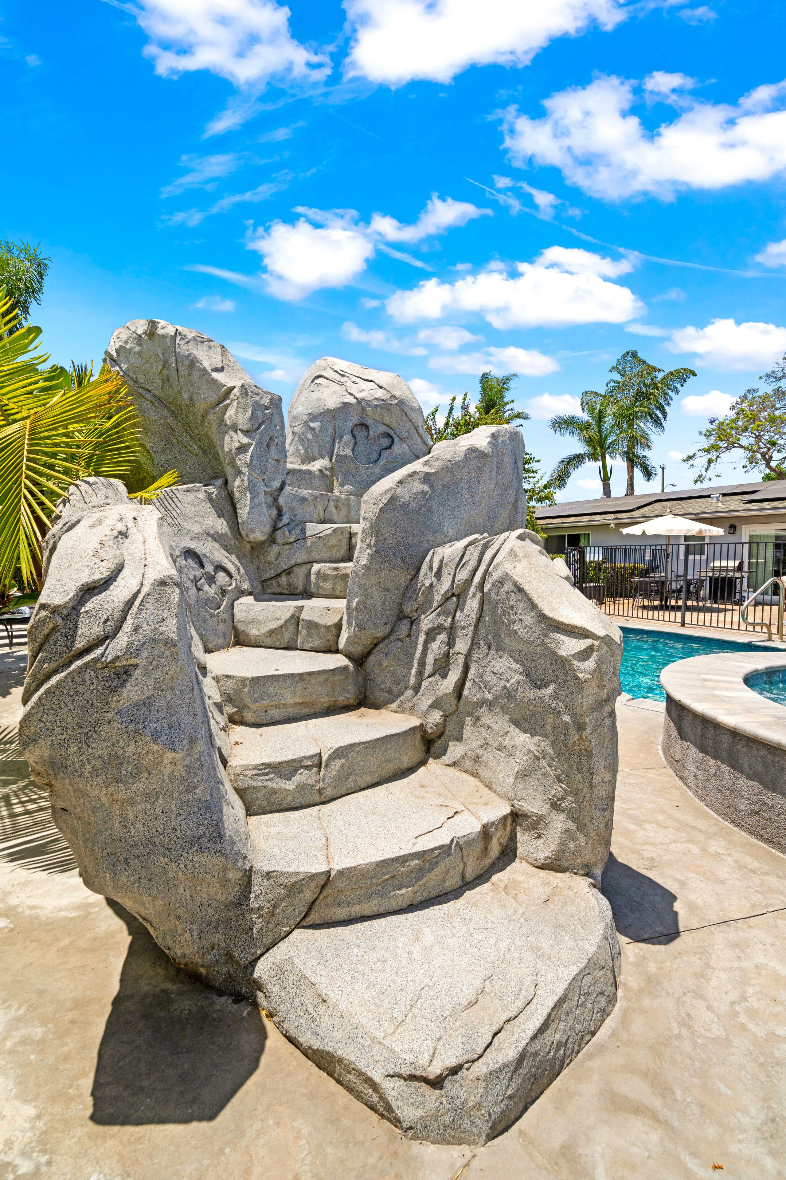 A stone rock formation with steps leading to a pool area, surrounded by palm trees and a clear blue sky.