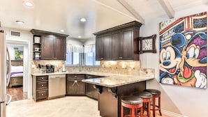 A modern kitchen with dark wood cabinets, a large island with stools, a clock, and a Mickey Mouse painting on the wall. - Beautiful 5bd 3ba Private Resort Pool and HotTub, Waterslide, CloseTo Disneyland (Anaheim)