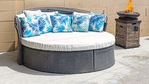 A round outdoor seating area with a white cushion, blue leaf-patterned pillows, and a stone fire feature. - Beautiful 5bd 3ba Private Resort Pool and HotTub, Waterslide, CloseTo Disneyland (Anaheim)