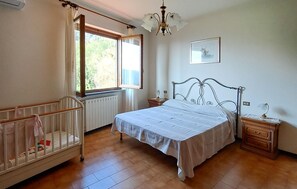 A bedroom with a bed, a crib, a window with curtains, a radiator, a bedside table, a lamp, and a framed picture on the wall.