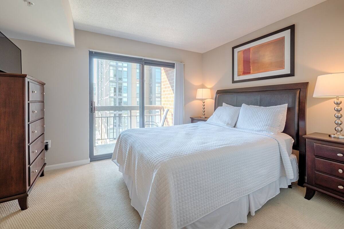 Une chambre à coucher avec un grand lit, une commode, une table de chevet, une lampe et une fenêtre donnant sur des immeubles.