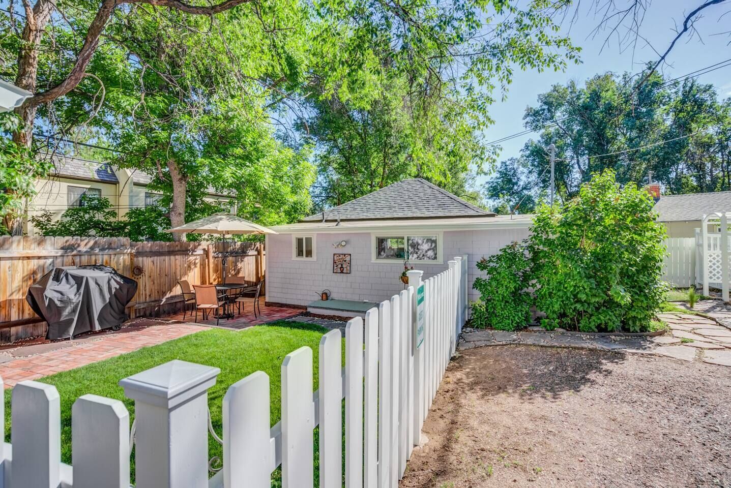 Ein Garten mit Terrasse, Grill, kleiner Gartenhütte und weißem Zaun.