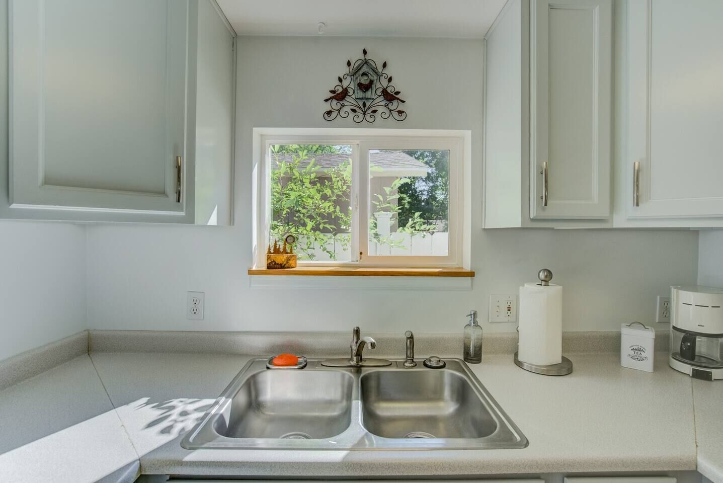 Una cocina con un lavamanos doble, una ventana que da a vistas de vegetación y un adorno metálico sobre la ventana.