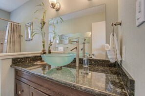 Un baño con lavamanos de vidrio, un espejo, un perchero para toallas y una planta.