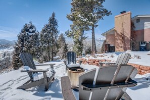 Un área de estar al aire libre cubierta de nieve con sillas Adirondack y un brasero, con vistas a un paisaje montañoso y un edificio de fachada de piedra.