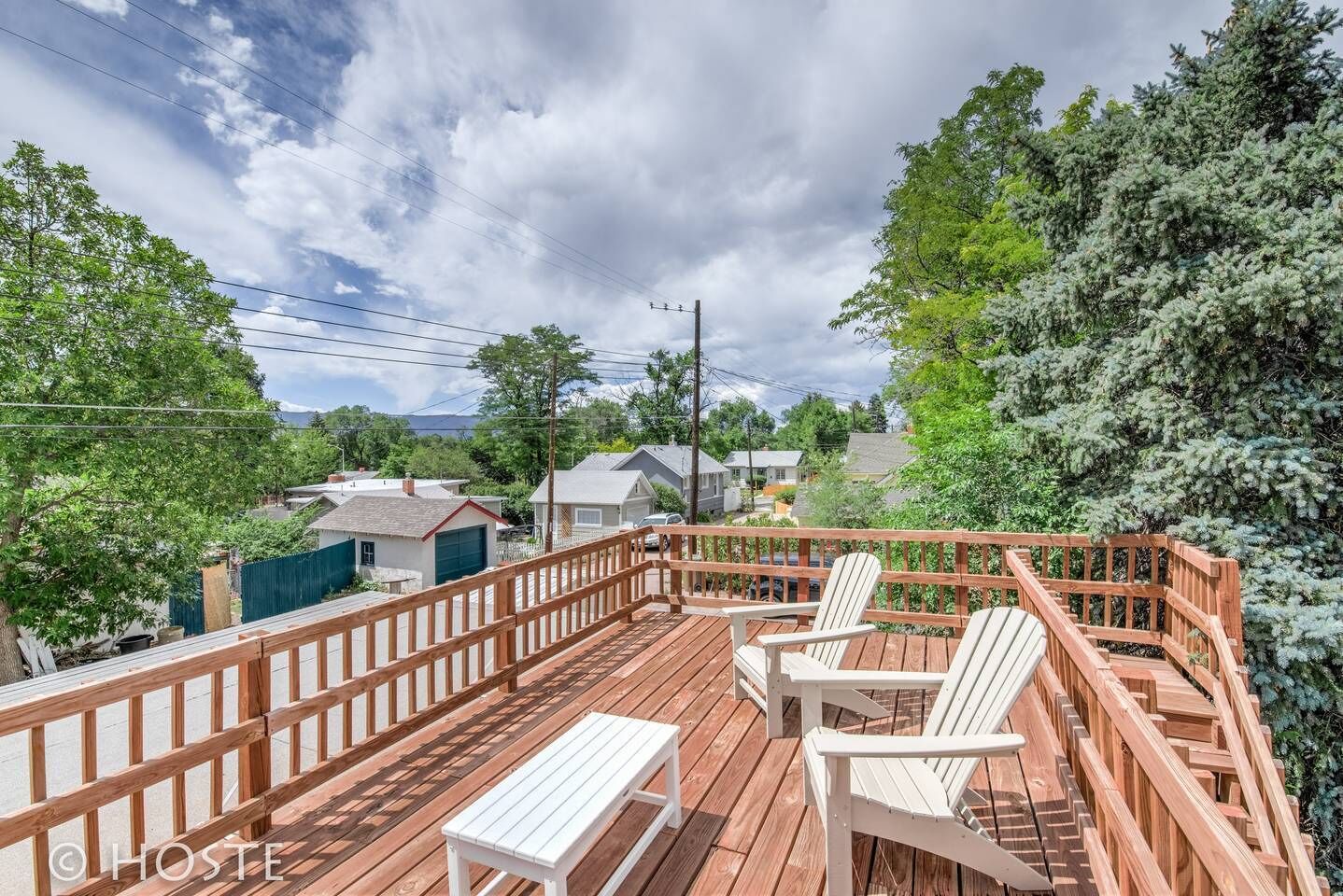 A wooden deck with white chairs and a bench, surrounded by a wooden railing, overlooking a residential area with houses and trees.