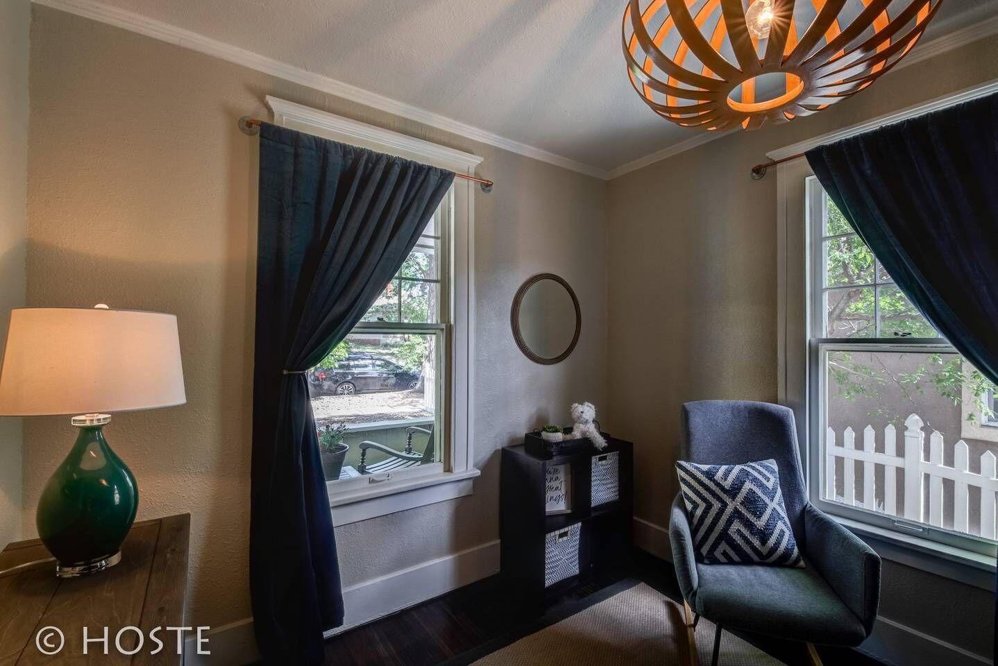 A room with two windows, a lamp, a chair, a small table, and a decorative mirror.